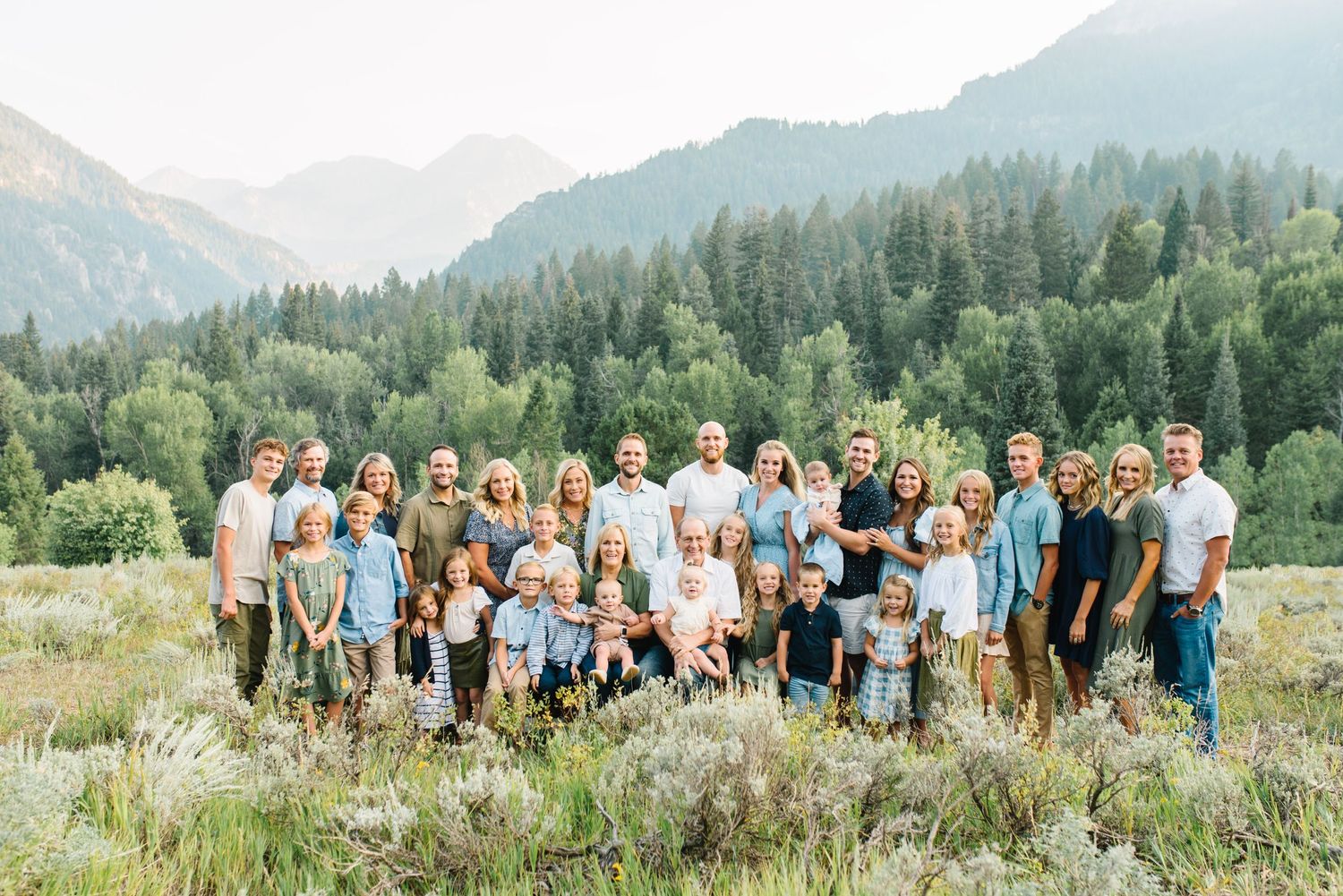 Extended family gathering poses in mountain meadow with evergreen forest backdrop.