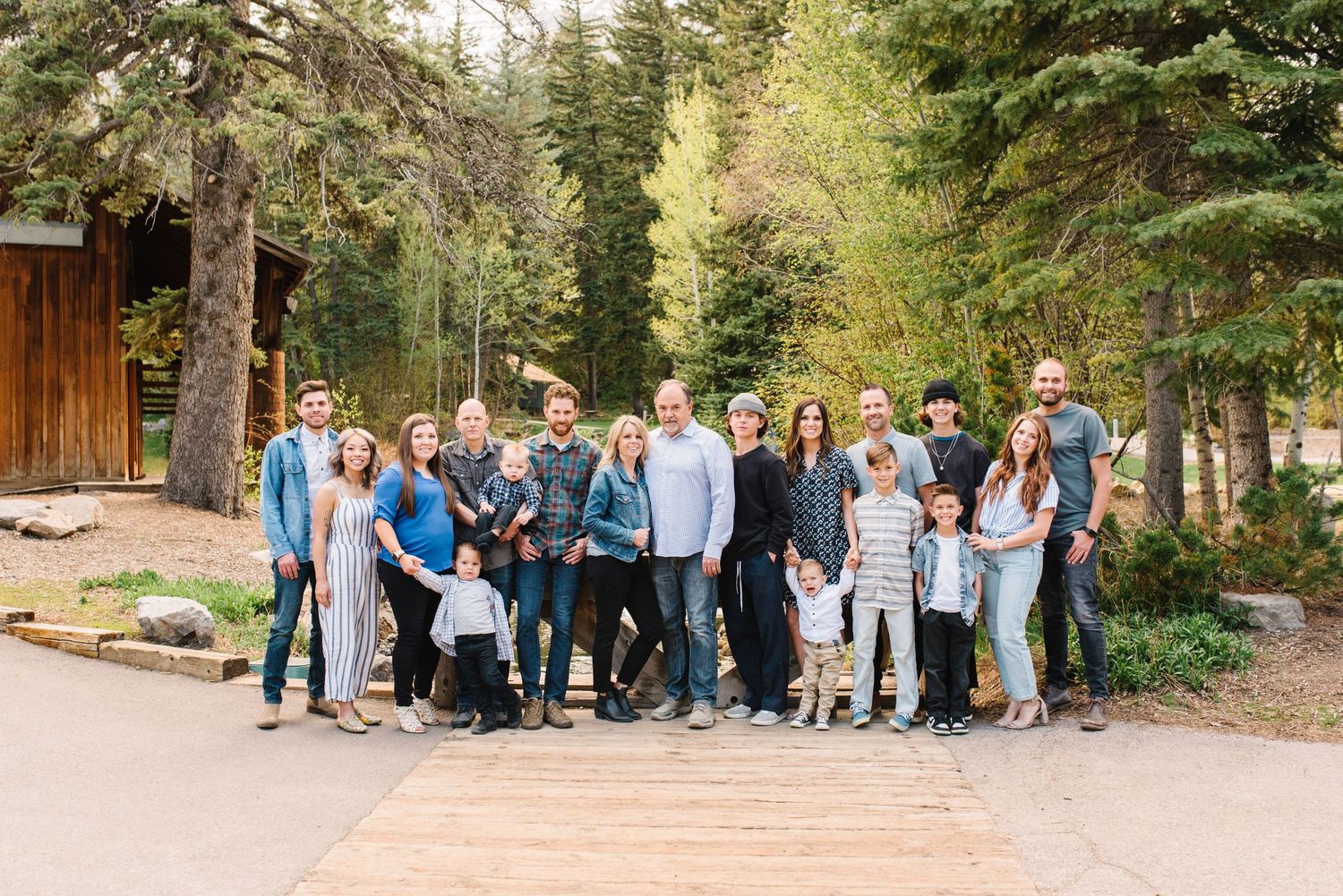 Large group gathered outdoors in casual attire on a wooded path with evergreen trees in the background.