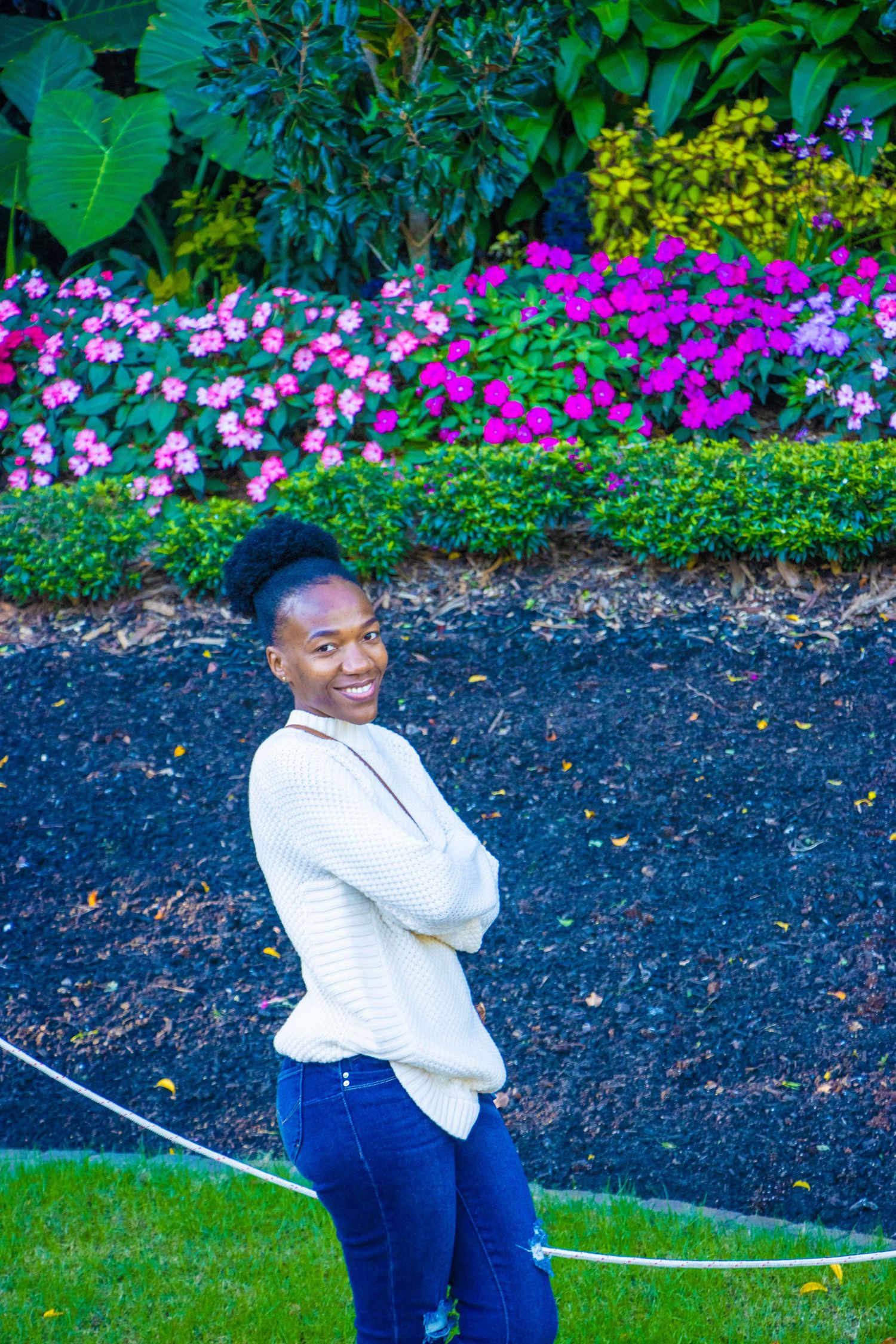 A person in a white sweater smiles while standing in front of a colorful garden with pink and purple flowers.