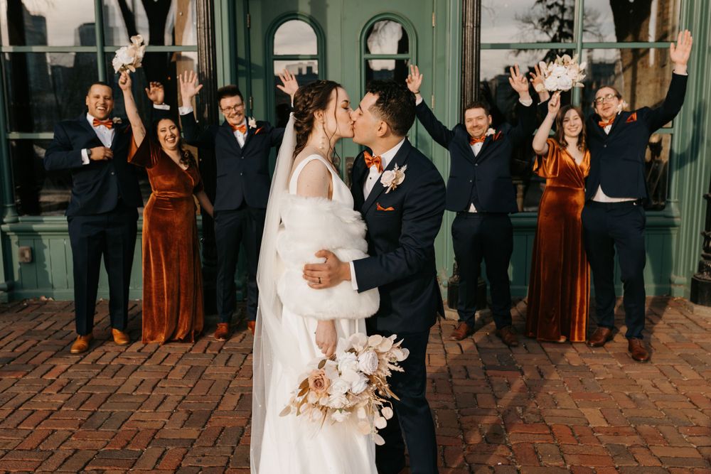 Bride and groom share a kiss while wedding party cheers with raised arms.