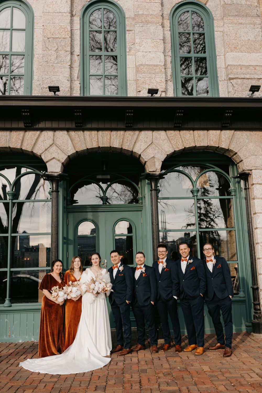 Bridal party stands before historic stone building with green windows in coordinated attire.