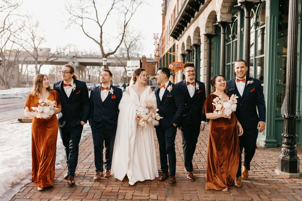Wedding party walks along brick sidewalk in rust velvet and navy attire.