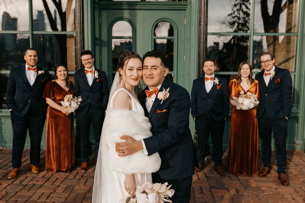 Bride and groom embrace while wedding party celebrates behind them outdoors.