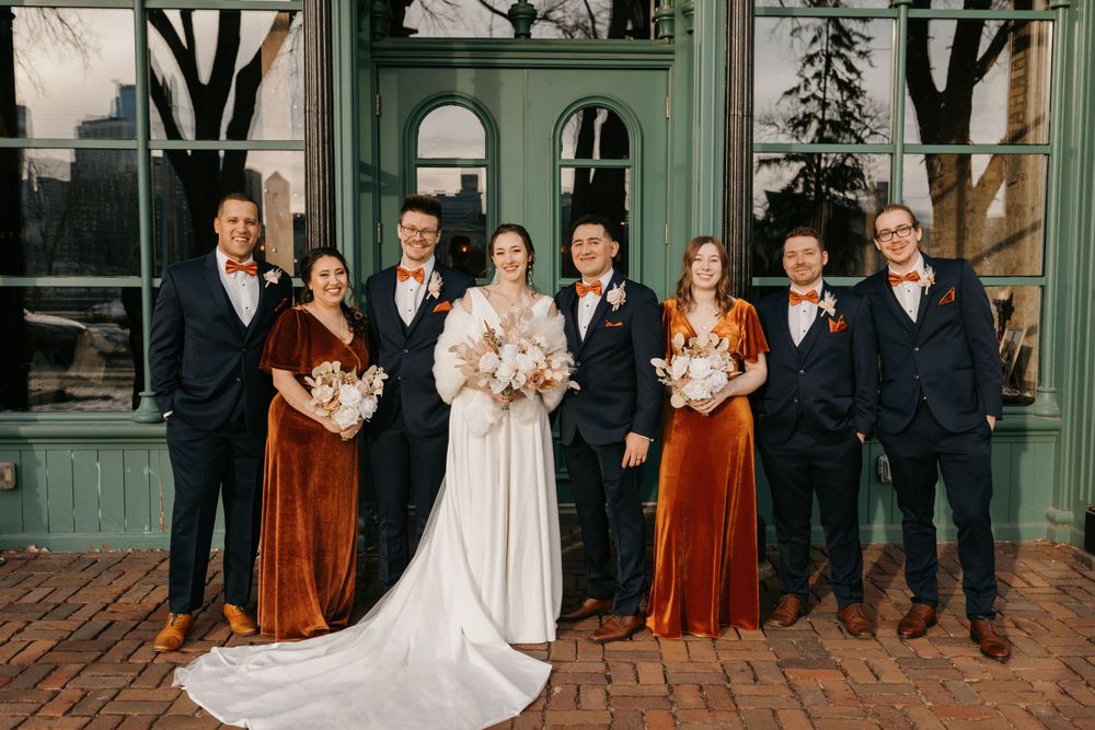 Wedding party poses in rust velvet dresses and navy suits before green building.