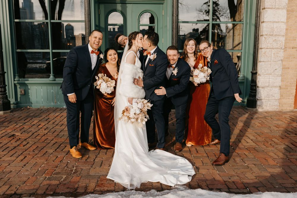 Wedding party surrounds kissing bride and groom on brick patio outside venue.