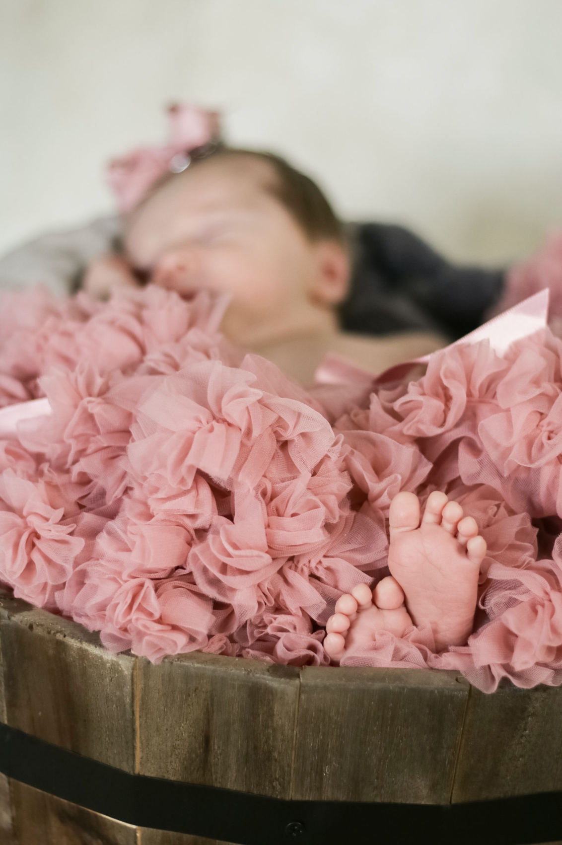 Sleeping newborn in wooden bowl surrounded by pink ruffled fabric.