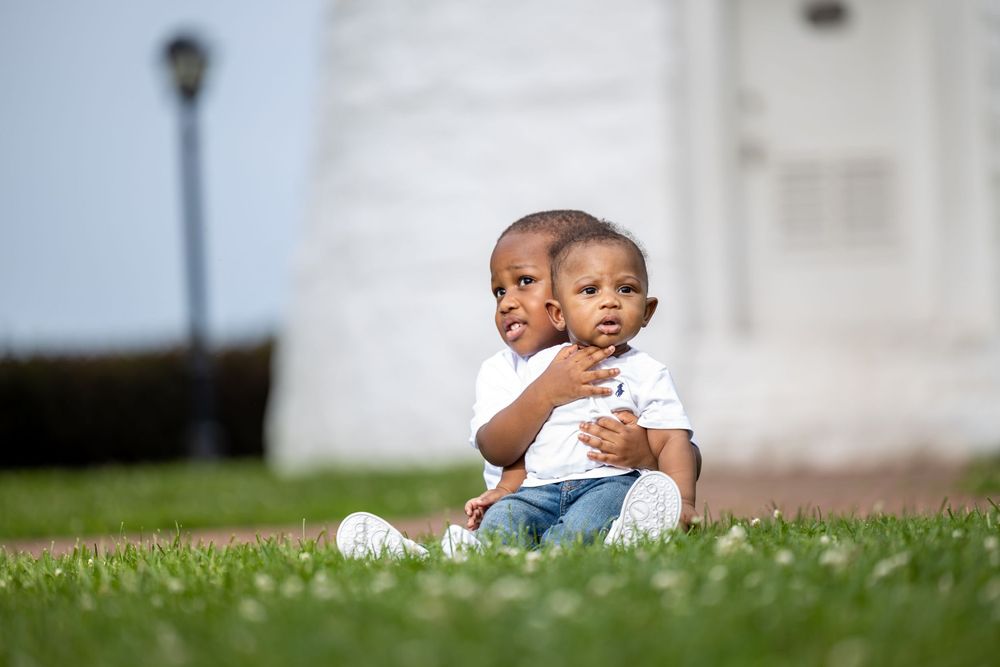 Two young children sit on grass in front of white lighthouse wall.