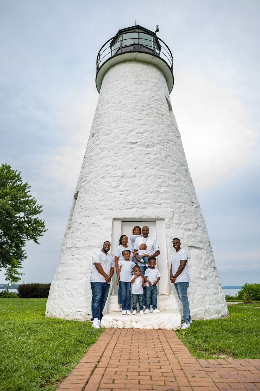 Family group poses in doorway of white lighthouse on sunny day with brick path leading to entrance.