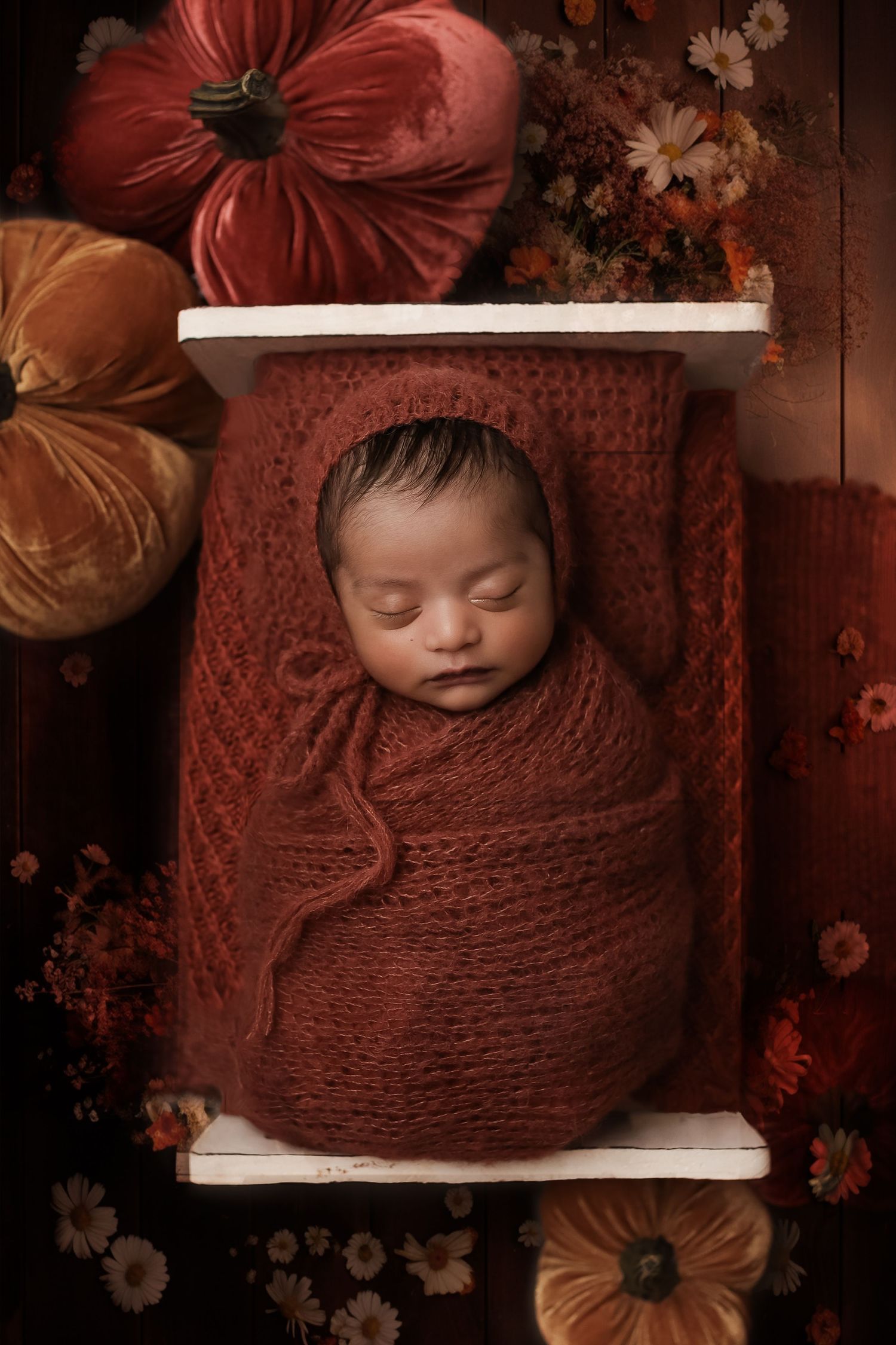 Sleeping newborn wrapped in rust-colored knit blanket surrounded by autumn flowers in rustic still life photography setup.
