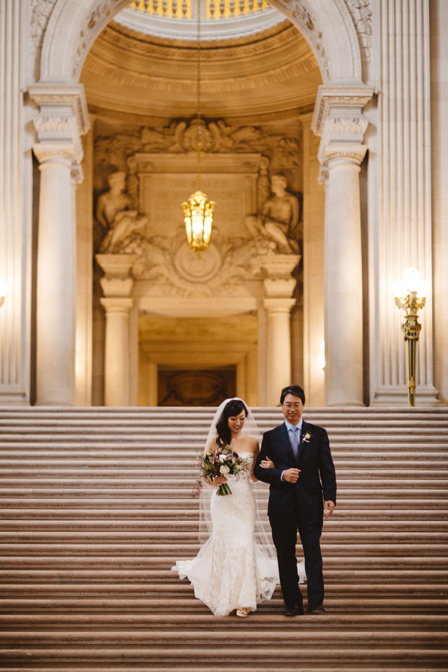 A couple descending ornate marble stairs beneath a decorated arch with classical statuary.