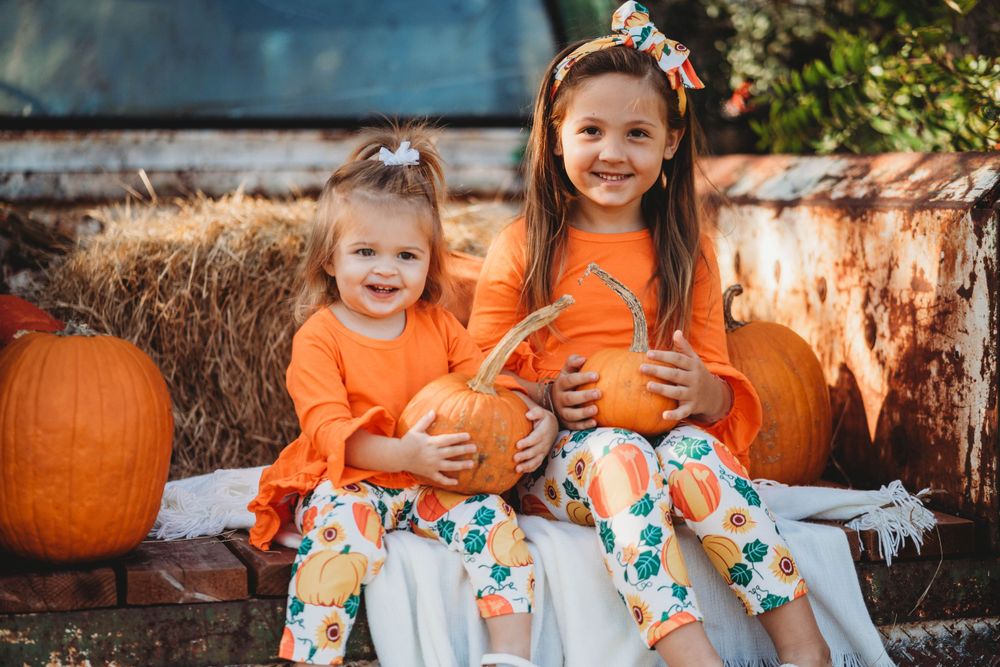 Children in orange outfits sit with pumpkins on rustic truck bed during fall photoshoot.