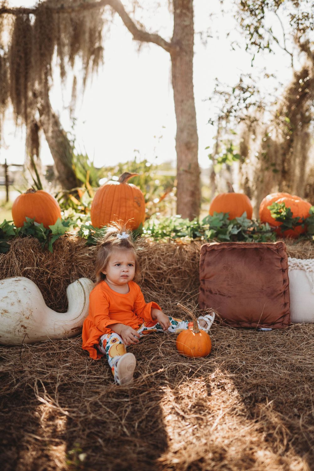 Child in orange dress sits among pumpkins and fall decor under moss-draped trees.