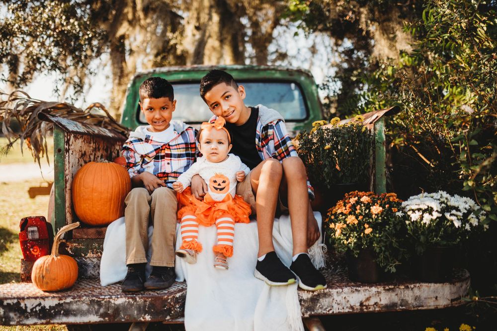Three children sit in back of vintage truck surrounded by autumn pumpkins and flowers at golden hour.