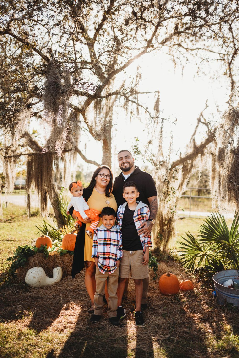 Family poses for fall photo with pumpkins and Spanish moss draped trees in background.
