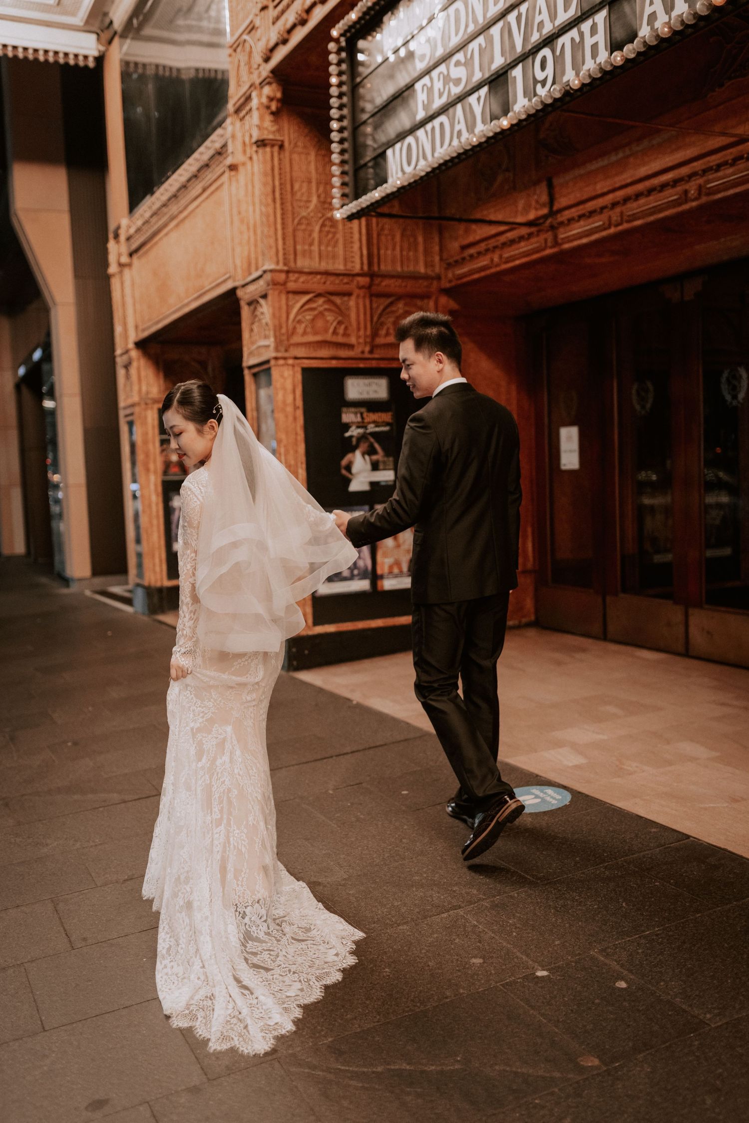 Bride in lace gown and groom in black suit pose outside a vintage theater marquee on their wedding day.
