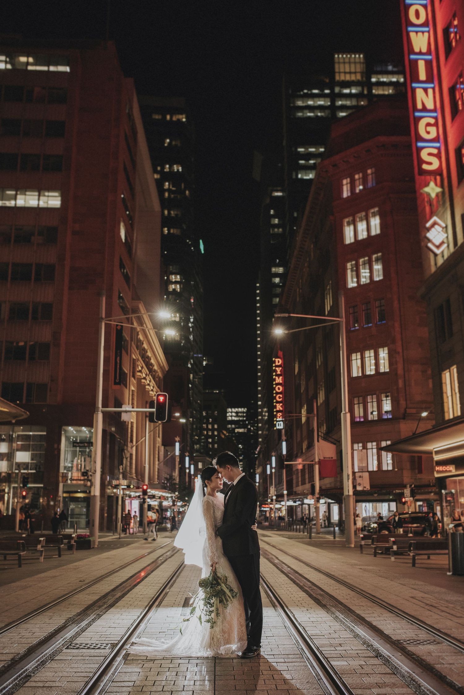 Bride and groom share a romantic kiss on city tram tracks surrounded by glowing urban nightlife.
