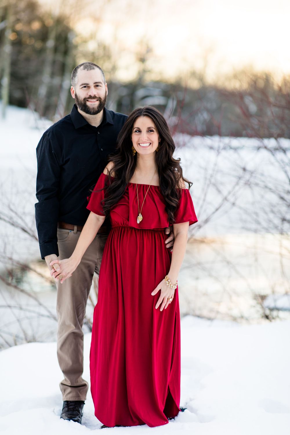 A couple poses together in the snow, with her in a flowing red dress and him in dark clothing.