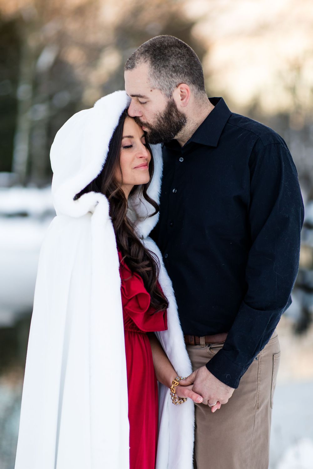 Couple embracing in winter scene with white cape and red dress against snowy backdrop.