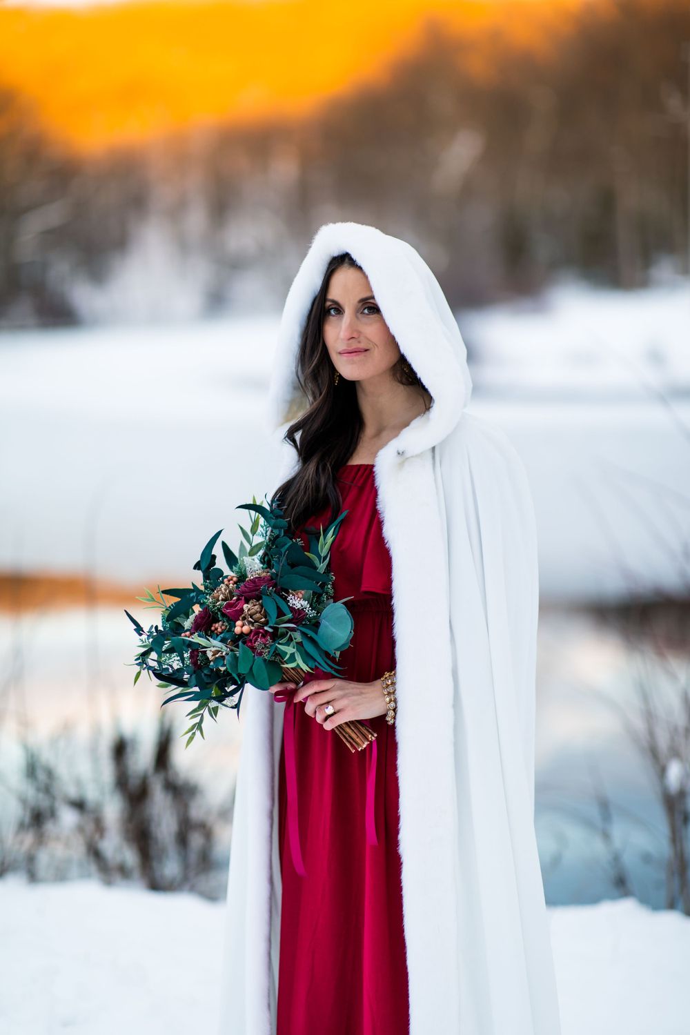 Woman in red dress and white cape holds greenery bouquet in snowy winter setting.
