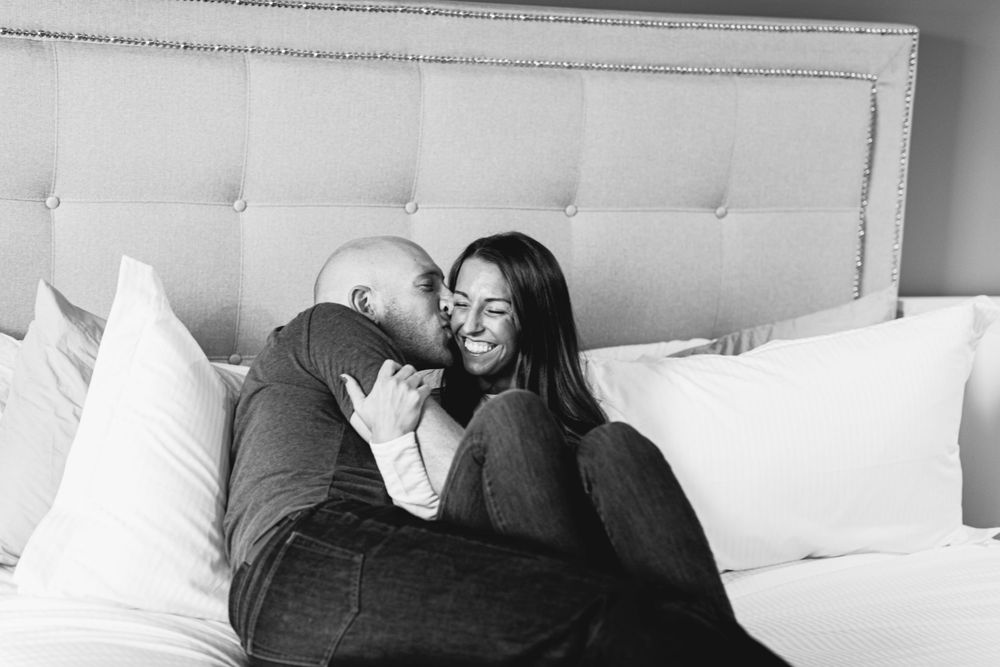 An intimate black and white photo of a couple sharing a tender moment on a bed with white pillows.