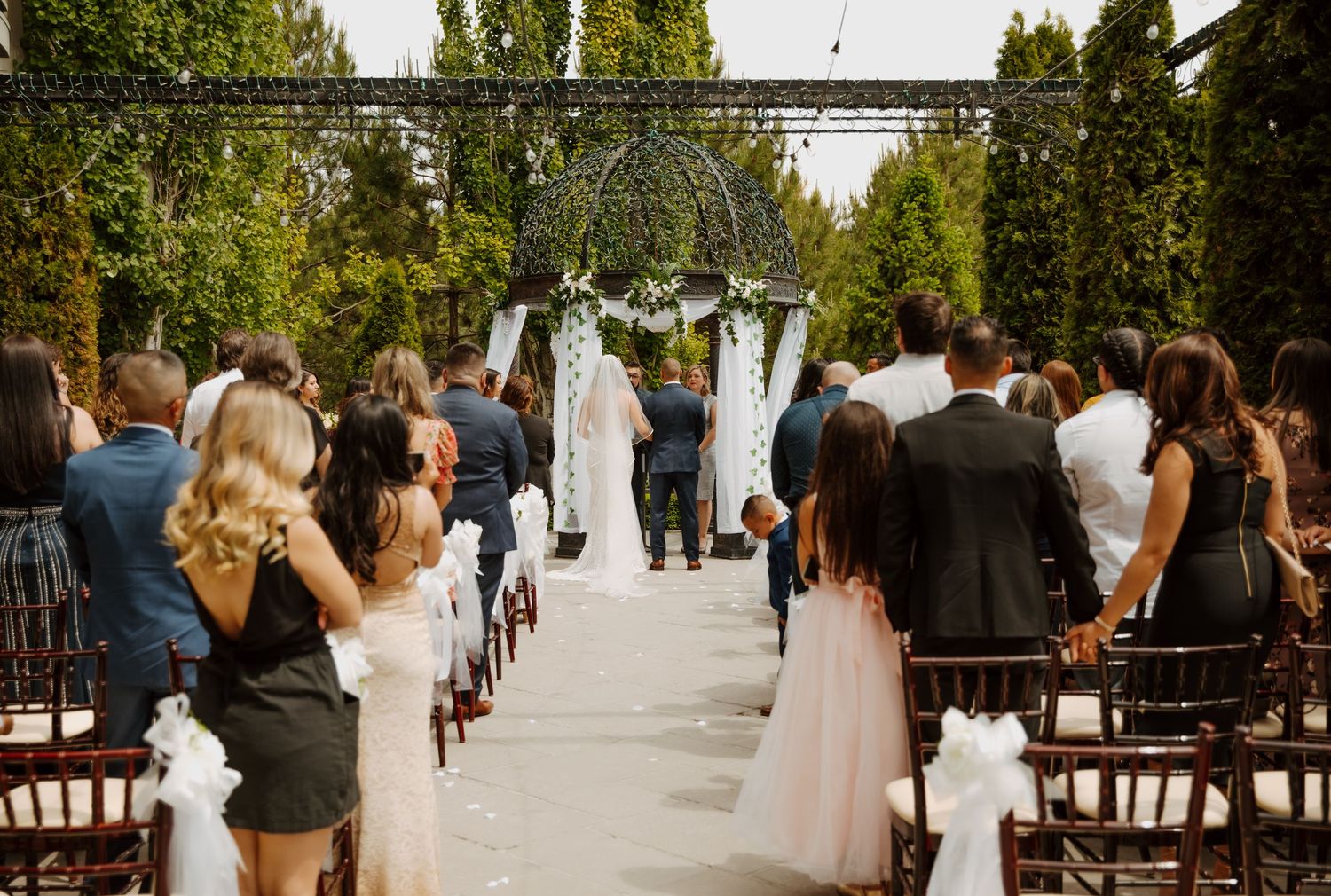 Outdoor wedding ceremony with white decor and rows of guests beneath tall trees.