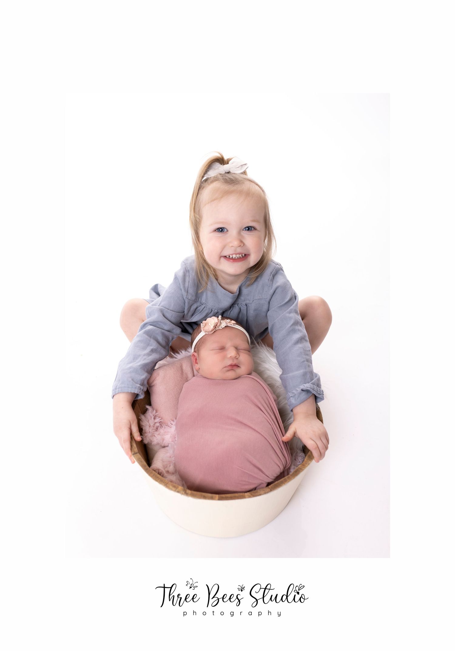 Siblings portrait with older child holding newborn in cream-colored bowl.