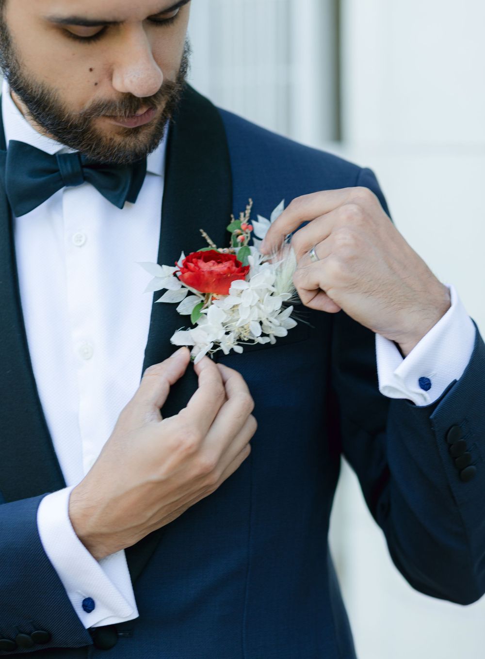 Close-up of groom adjusting red and white boutonniere on navy blue tuxedo lapel.
