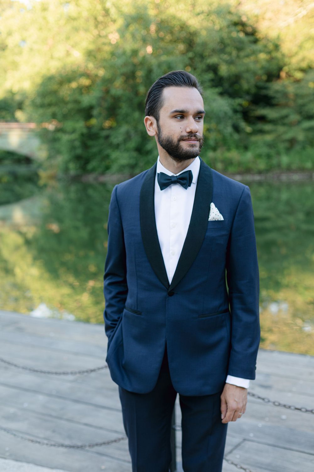 Groom in navy tuxedo and black bow tie poses by lake in natural park setting.