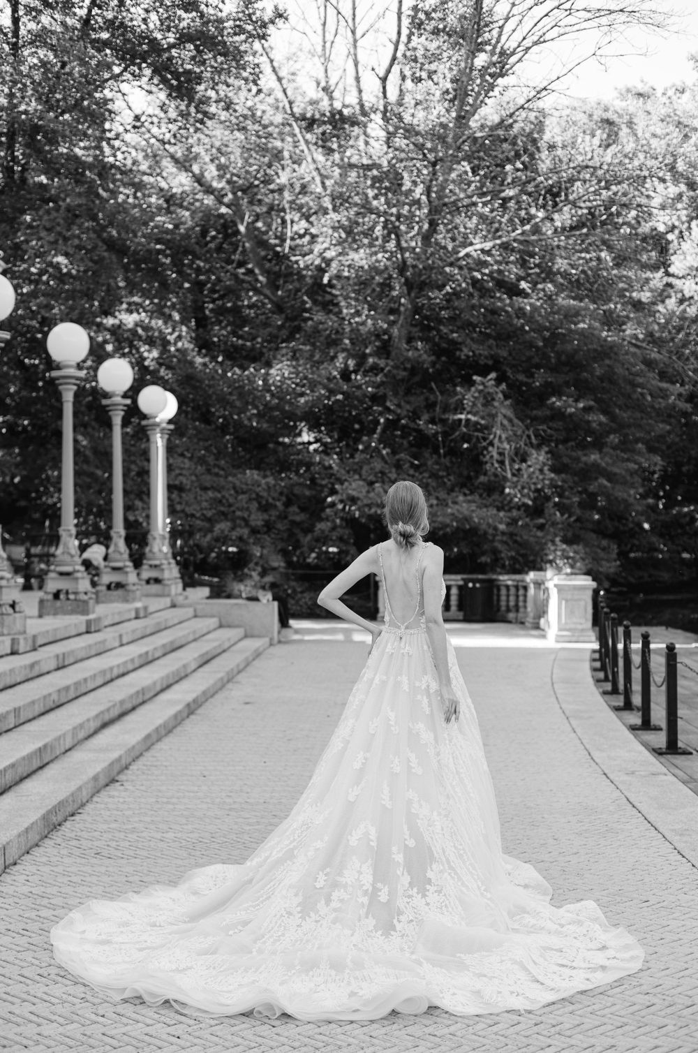 A flowing wedding gown with long train photographed from behind on stone steps with vintage lampposts.