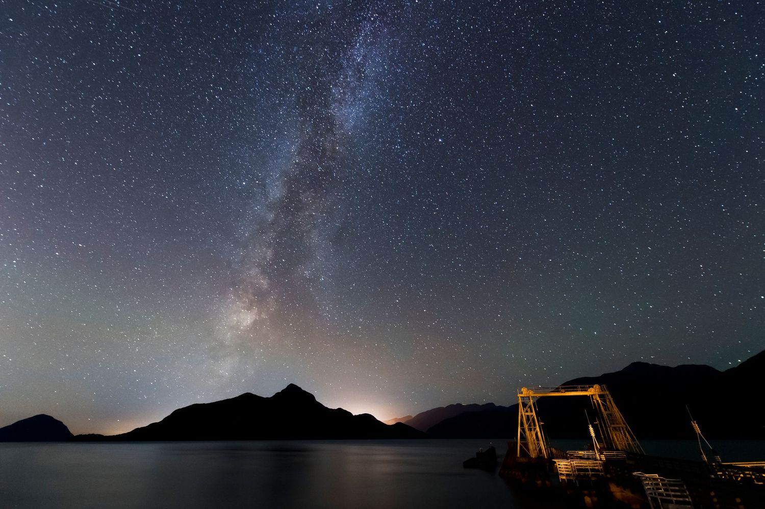 Milky Way galaxy stretches across night sky over mountain lake with camping equipment in silhouette.