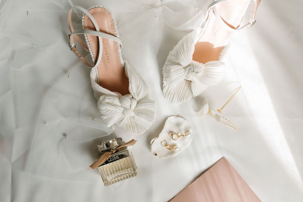 Elegant white bridal shoes and perfume bottle arranged on white fabric.