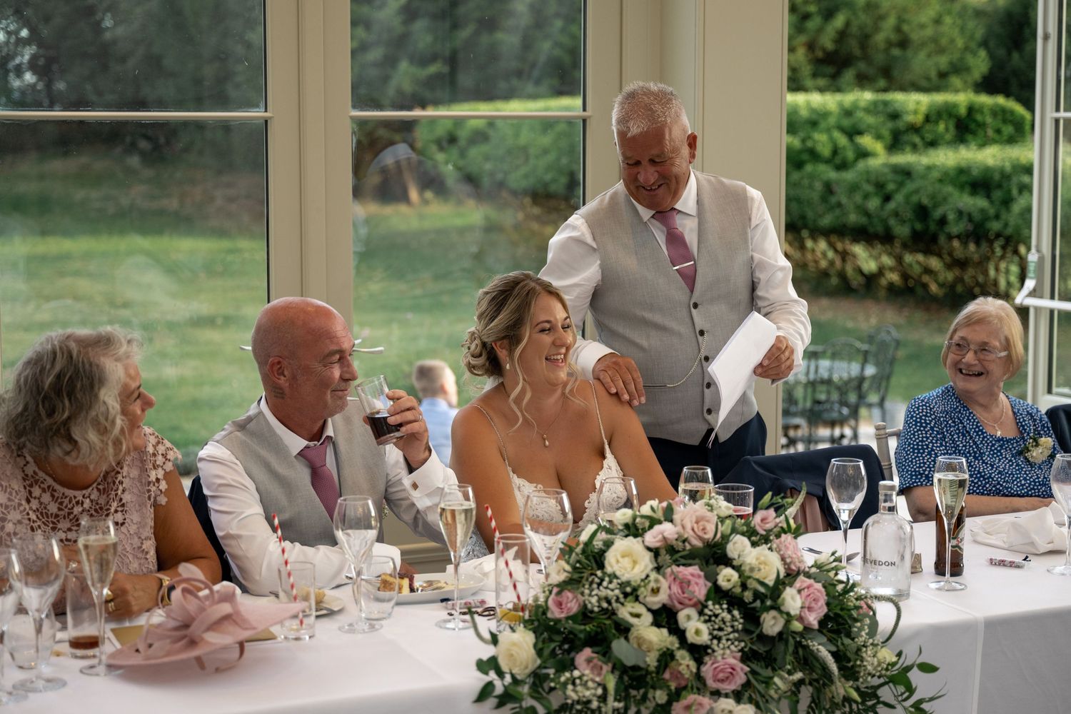 Wedding reception scene with guests seated at decorated table with pink floral centerpiece.