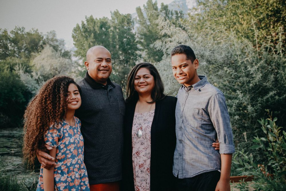 A family of four smiles together outdoors, posing in front of lush green trees and foliage.