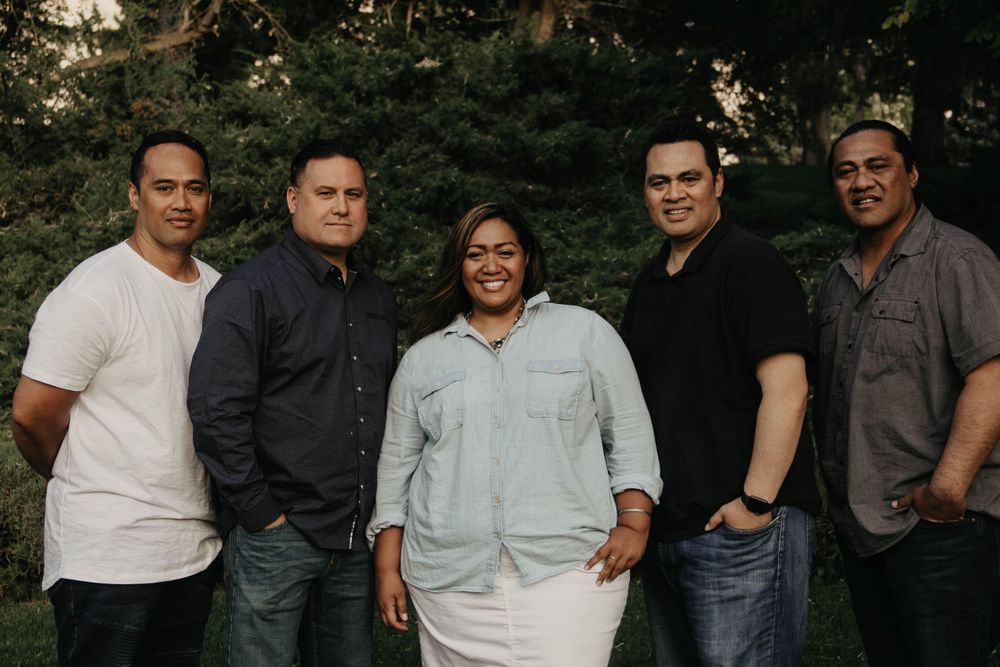 Five adults posing together outdoors in front of green trees for a family portrait.