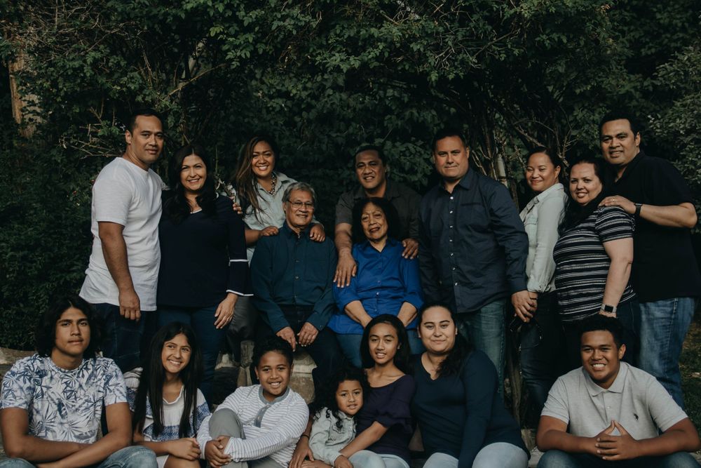 Large multi-generational family group portrait seated and standing outdoors near lush trees.