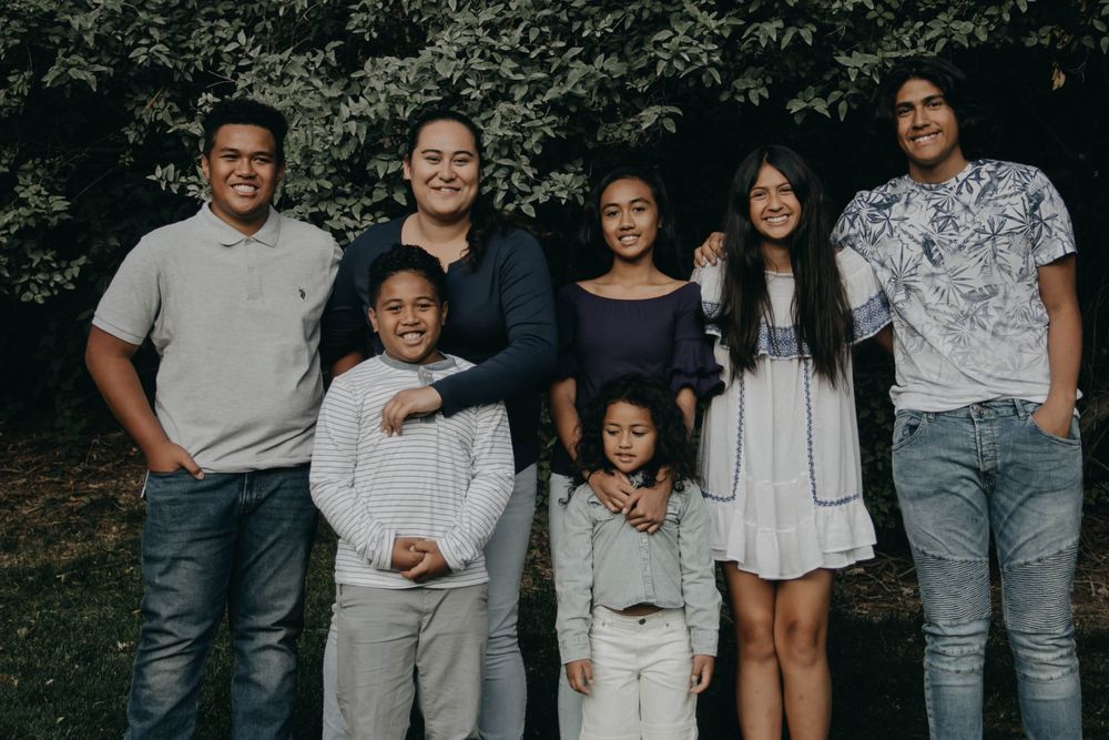 A large family of seven poses together outdoors, smiling warmly in front of lush green trees.