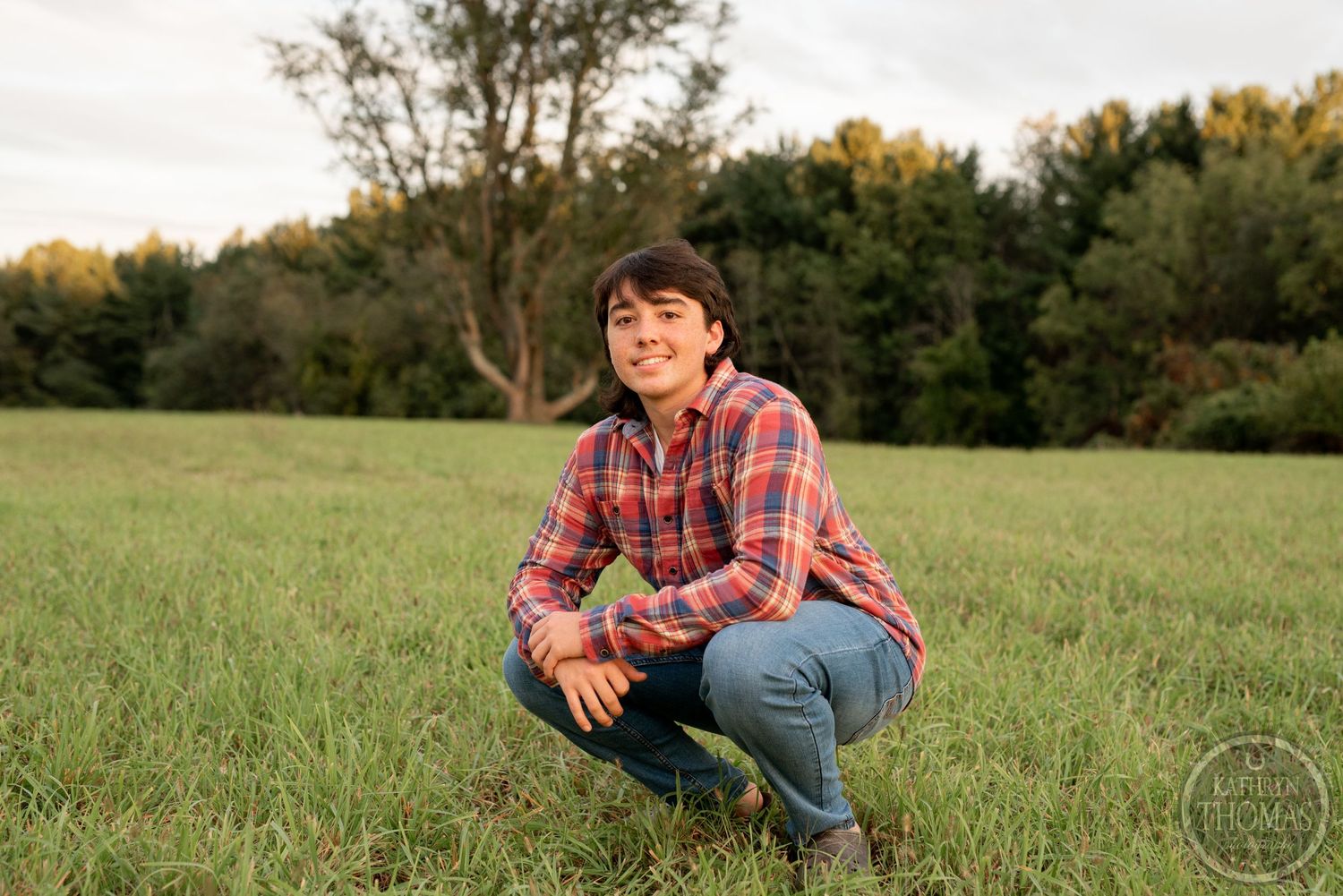 Person in plaid shirt crouches in grassy field during sunset.