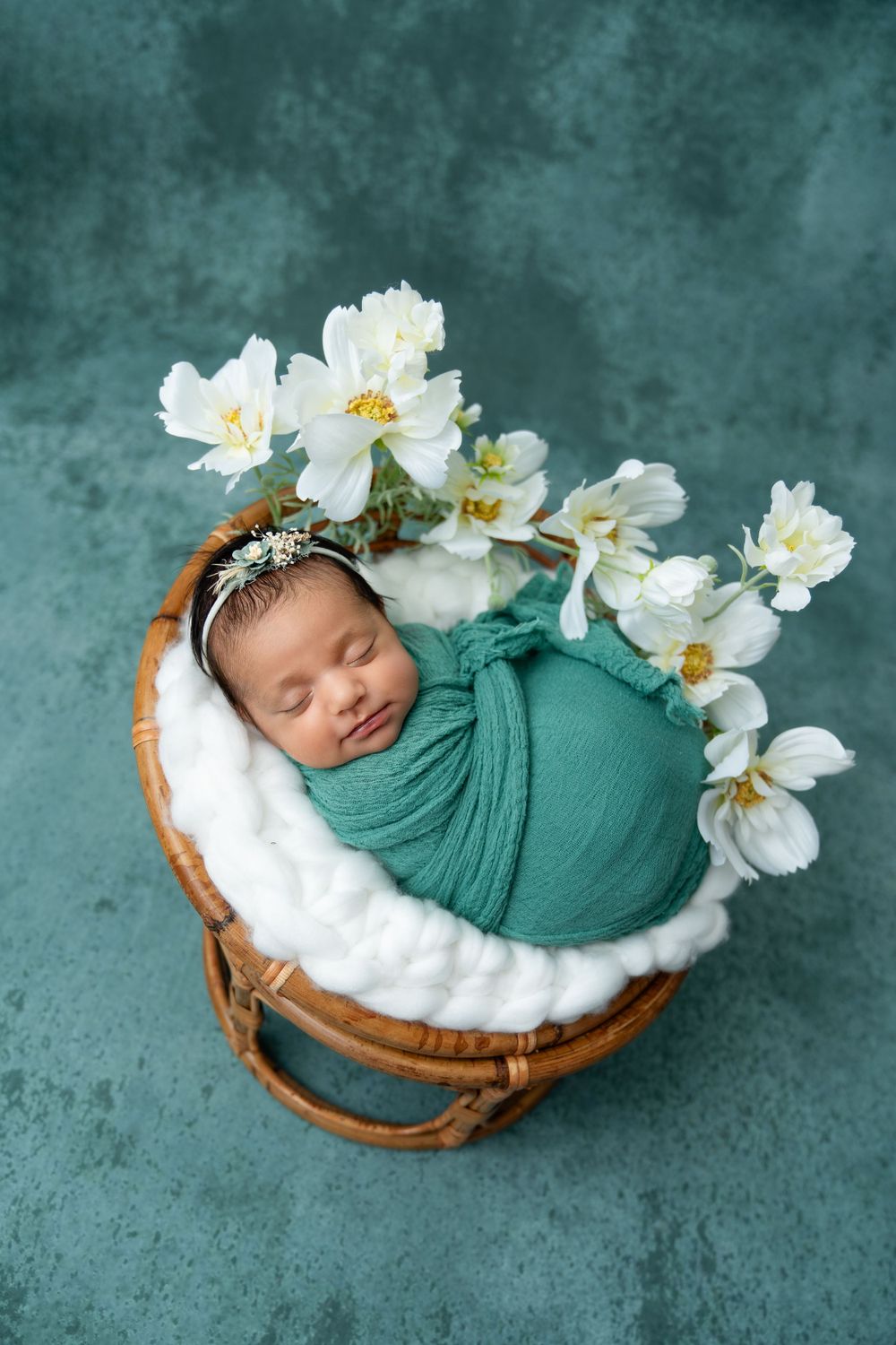 Newborn photography session with white orchids and teal wrap in wooden bowl against teal backdrop.