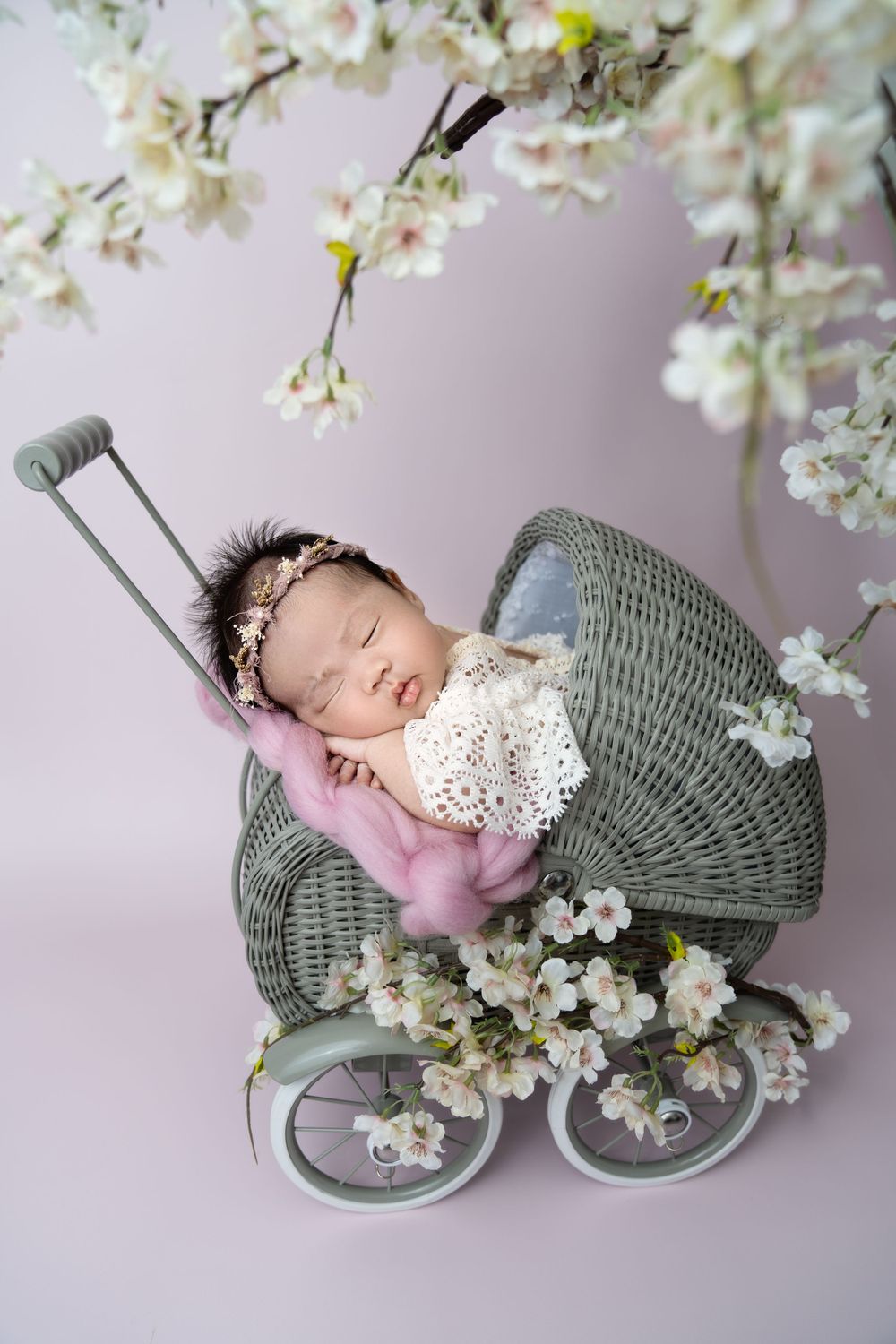A sleeping baby nestled in a grey knit basket surrounded by delicate white cherry blossoms.