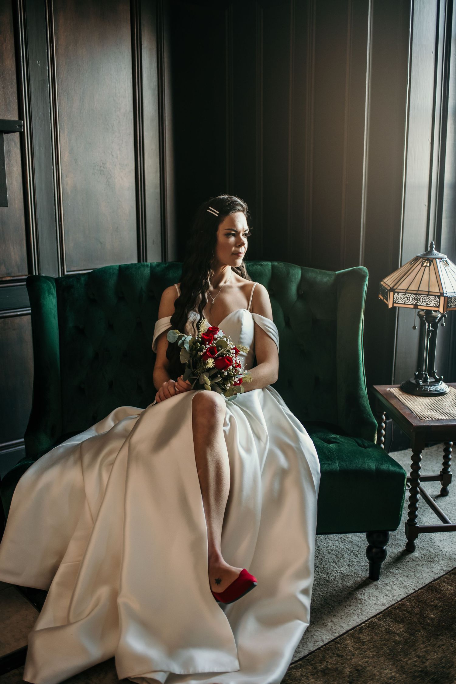 Bride in white gown sits elegantly on green velvet couch in dark moody room.