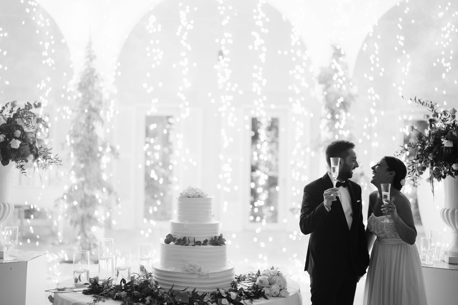 Newlyweds share a moment together next to their elegant white wedding cake with cascading fountain decorations.
