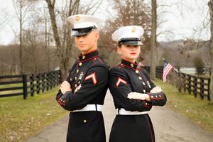 Two Marines in dress uniforms stand back-to-back on a path with wooden fences.
