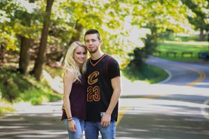 A couple poses together on a winding tree-lined road during a sunny day.