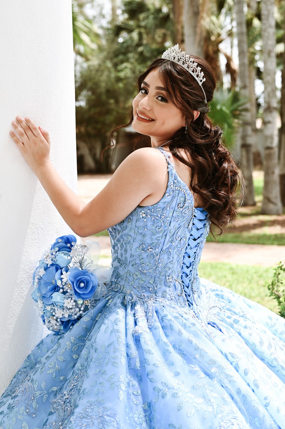 Beautiful blue quinceañera dress with sparkling tiara and bouquet against white column.