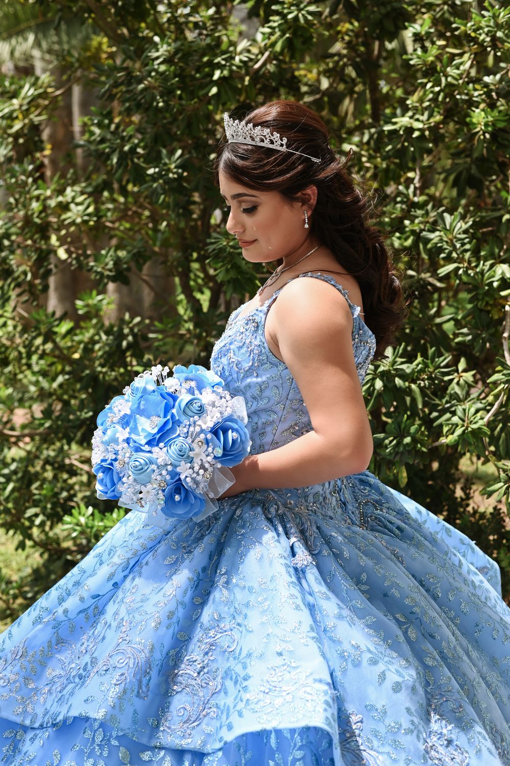 Light blue quinceañera gown with crystal tiara and blue bouquet in garden setting.
