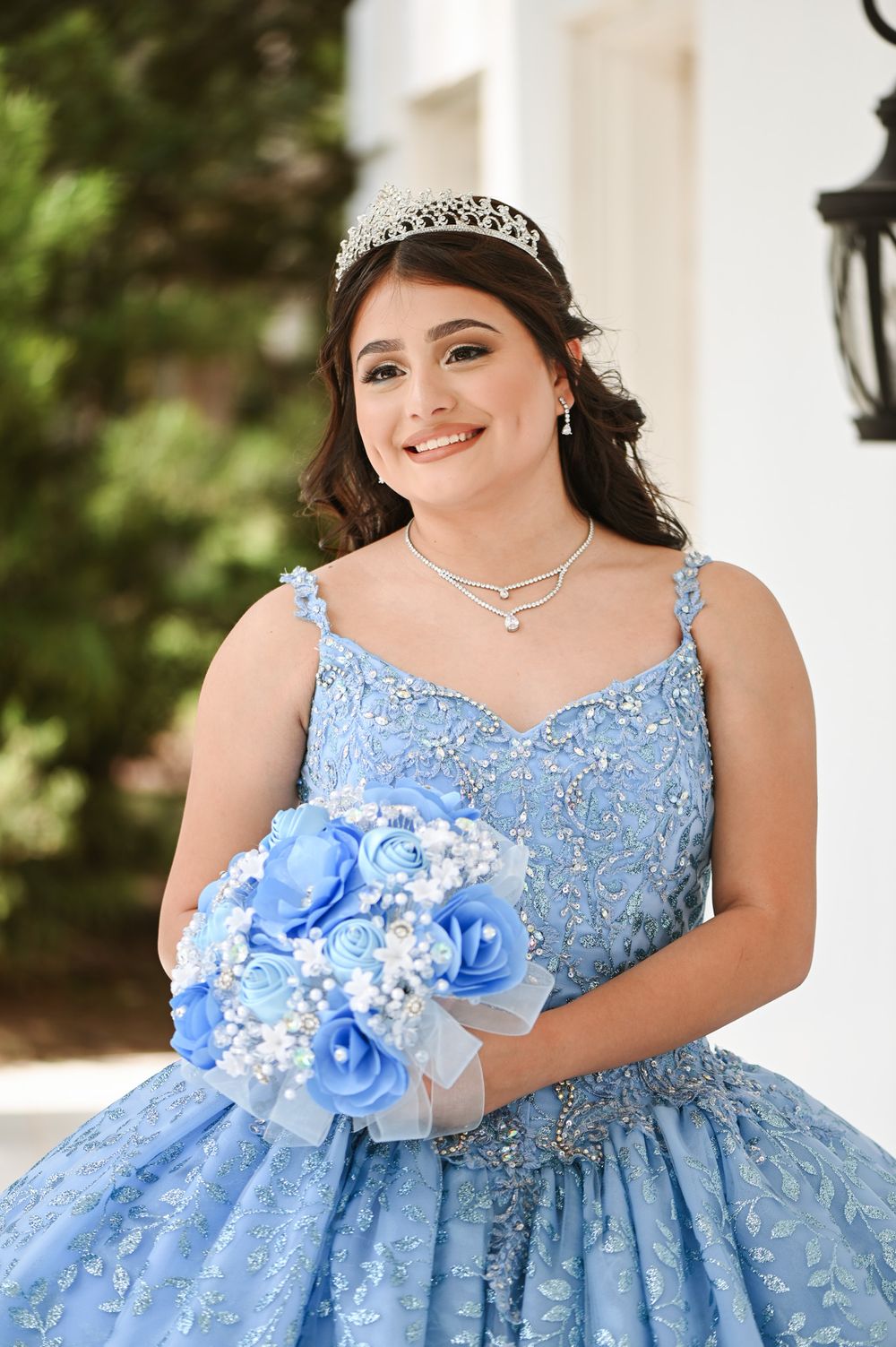 Elegant quinceanera dress in light blue with beaded details and matching blue bouquet against white backdrop.