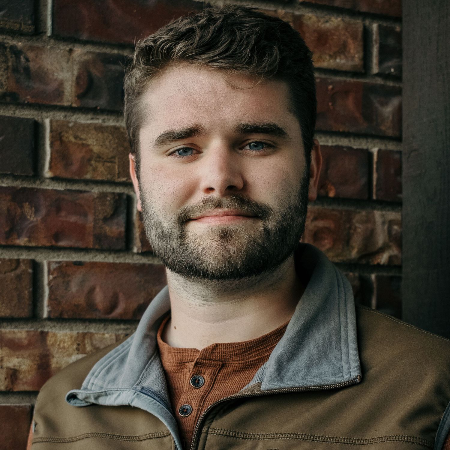 A close-up portrait against a brick wall wearing casual layers and sporting a neat beard.