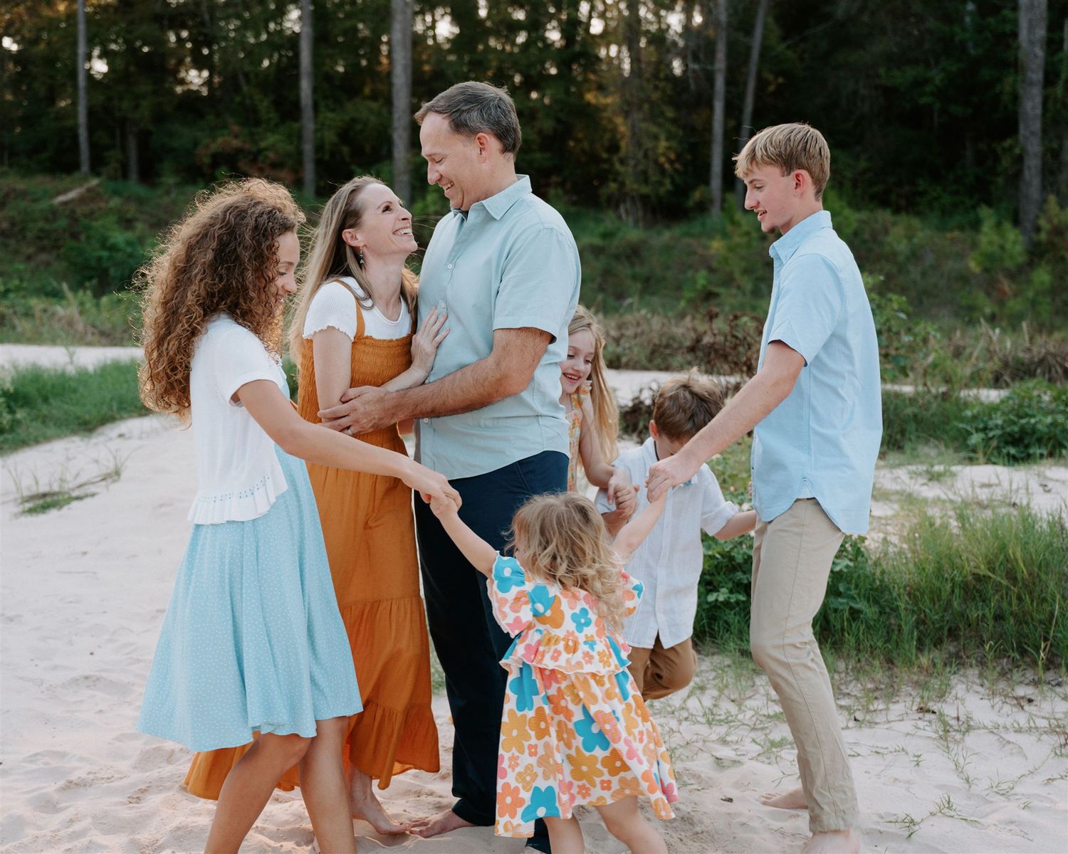 Family laughing and dancing together on sandy path with forest backdrop.