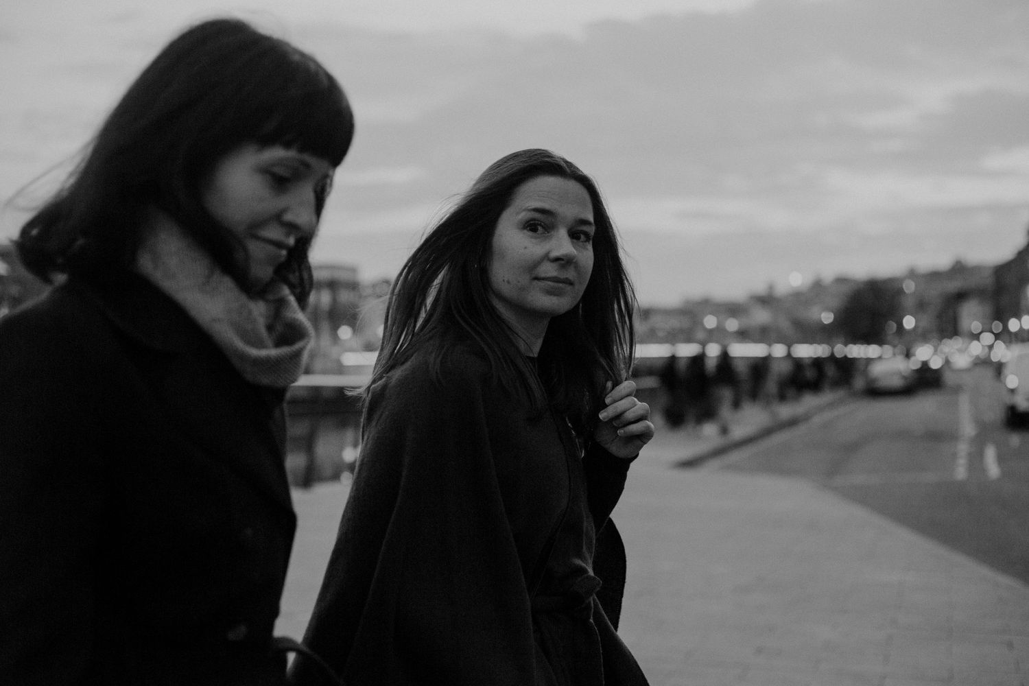 Two women in black coats stand together outdoors on an overcast day in black and white.