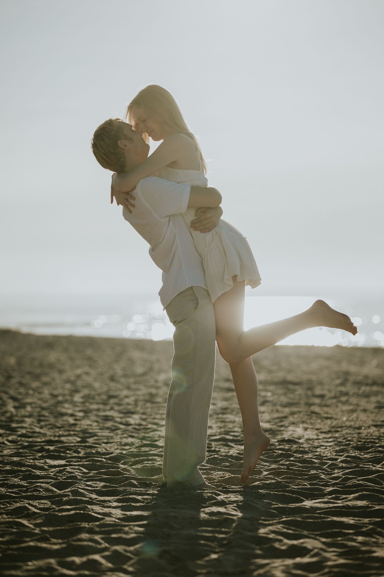 Guy lifts up his girl while nose to nose in front of the beach at sunset